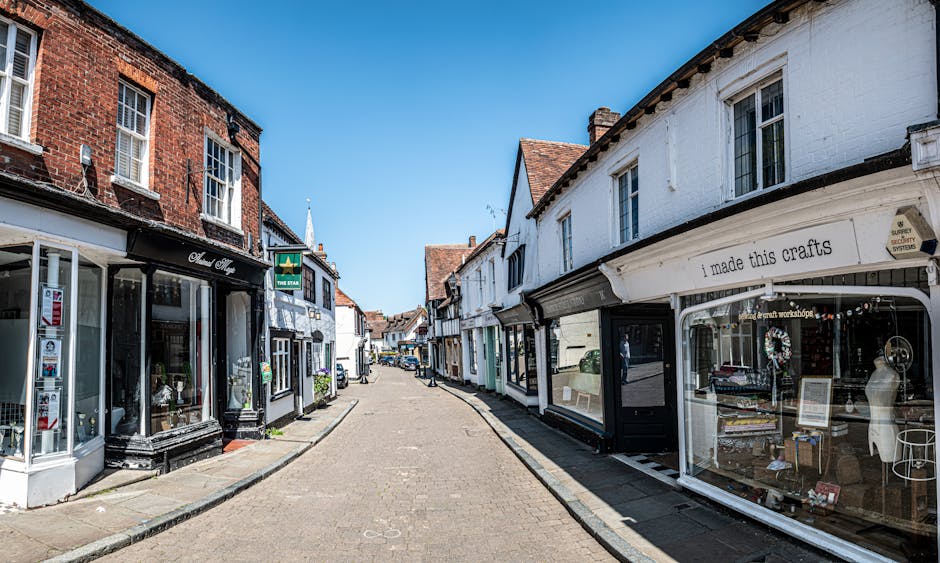 A wide street view of a small town high street with cobblestone paving, featuring storefronts on both sides including a craft shop displaying handmade items and signs, and a shop window with mannequin