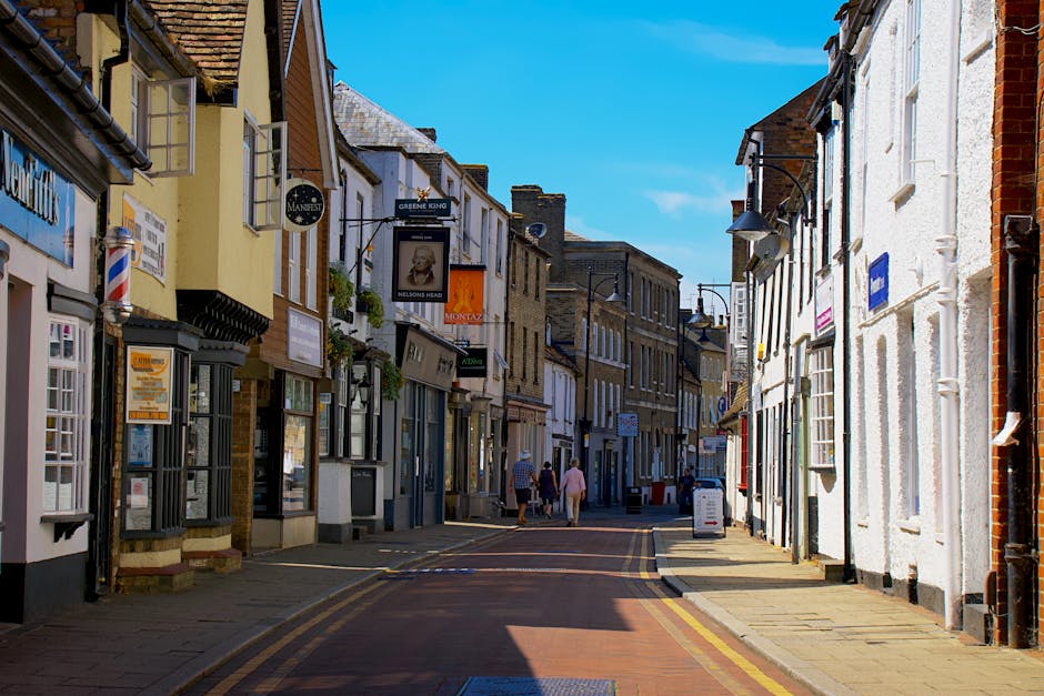 A wide street view of a small town high street with cobblestone paving, featuring storefronts on both sides including a craft shop displaying handmade items and signs, and a shop window with mannequin